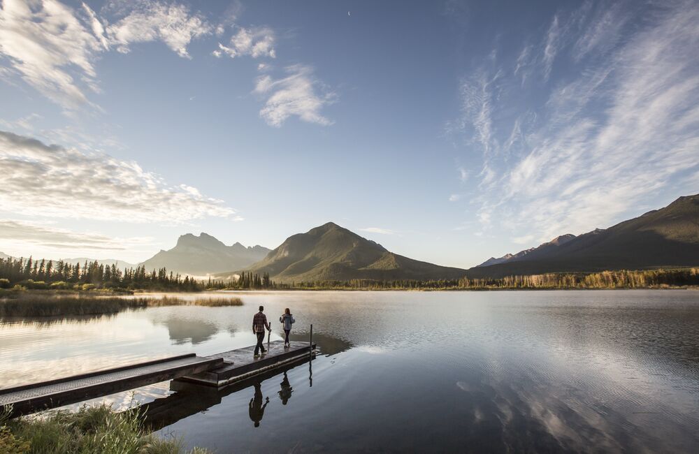 A couple walks along a dock towards a vast lake framed by Mount Rundle and Sulphur Mountain in Banff National Park.
